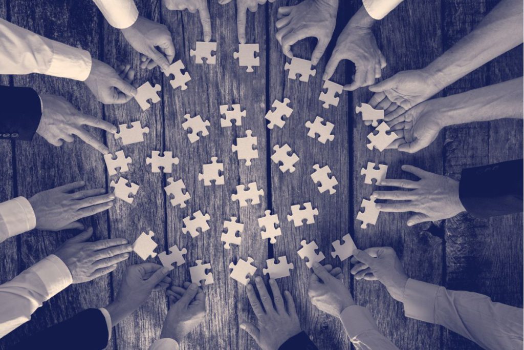 A group of people, their hands are outstretched across a tame in a circle, assembling puzzle pieces.