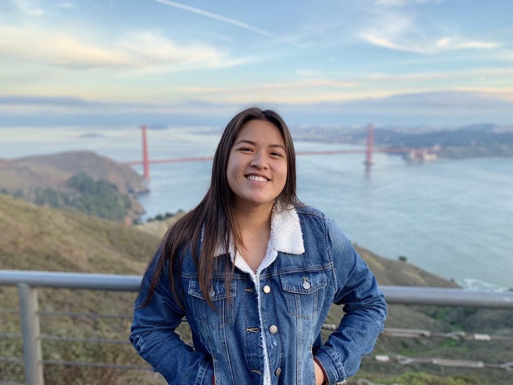 Han is standing in front of the Golden Gate Bridge. She is wearing a jean jacket and has her hands in her pockets.